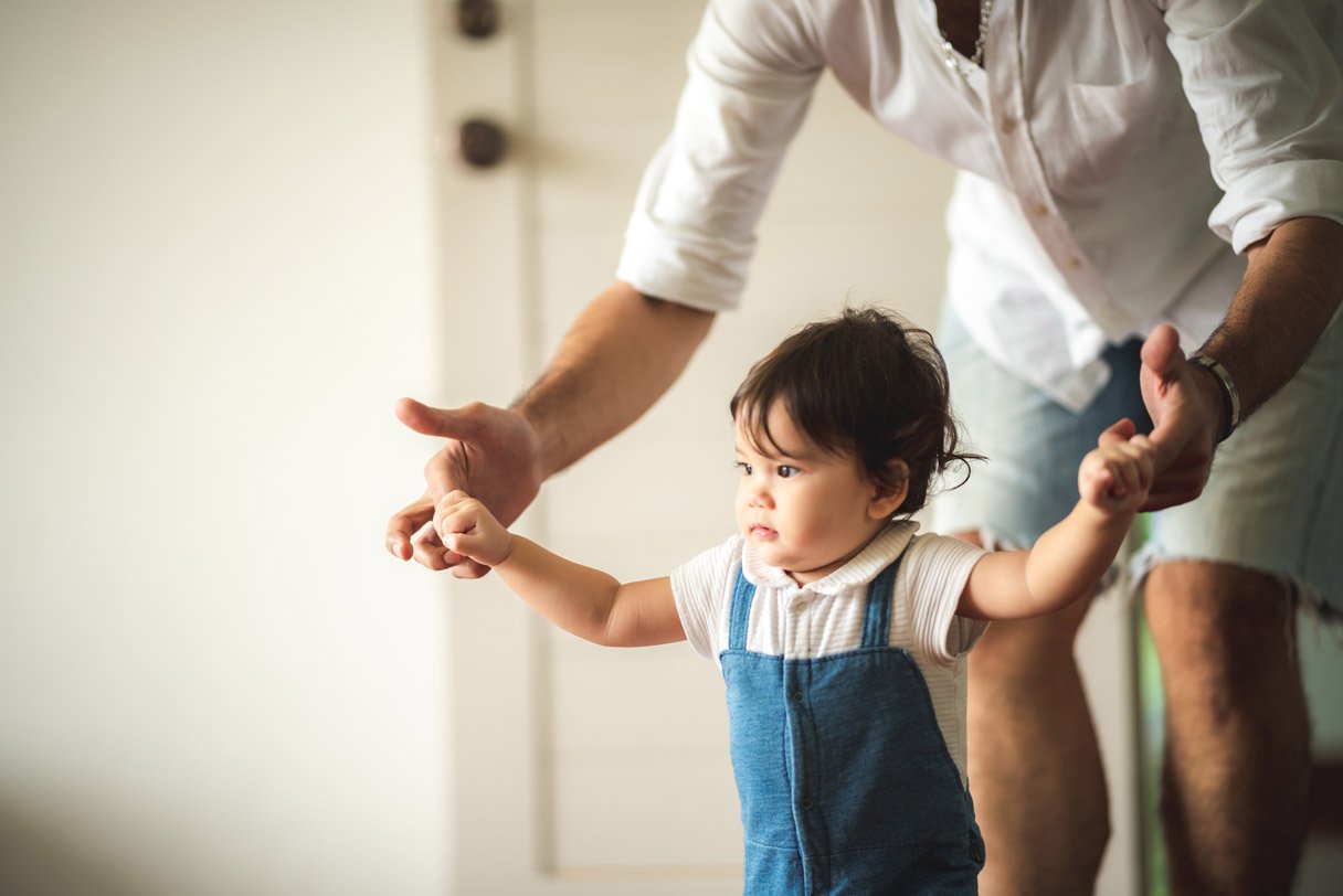 When do babies start walking - a toddler taking first steps while holding a parent's hands